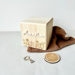 May include: A wooden box with a slot on top, engraved with the name "Aurelia" and a floral design. The box comes with a key and a small wooden circle with the text "frilly designs".