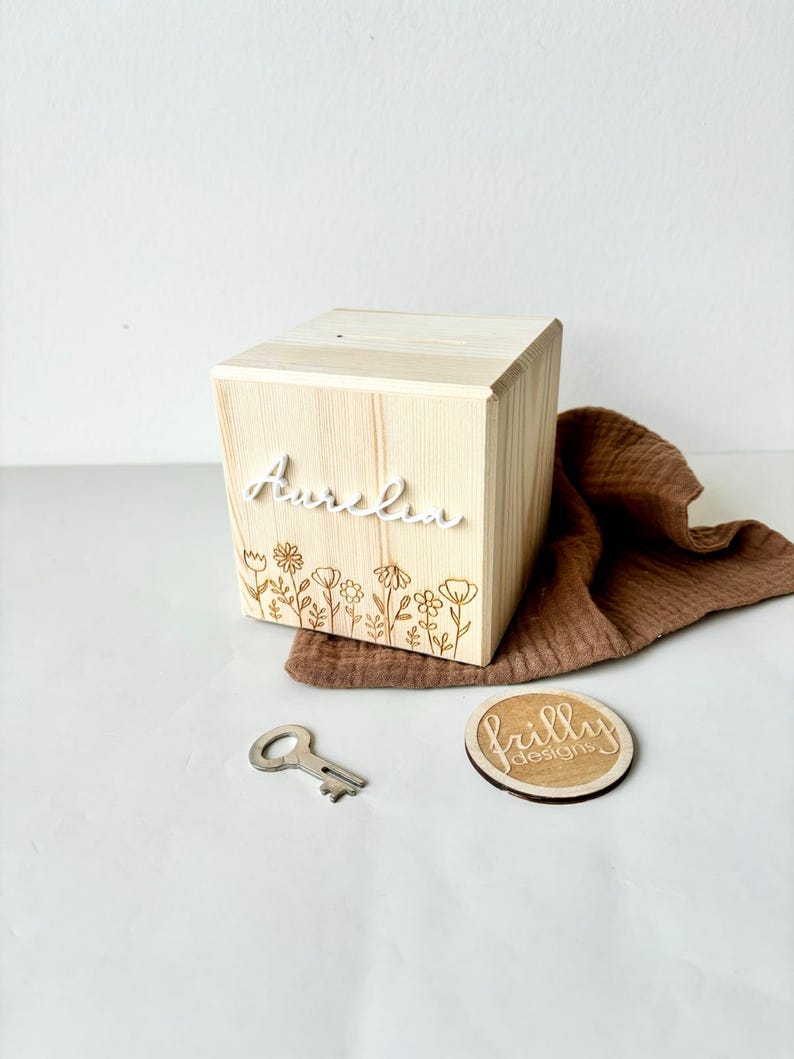 May include: A wooden box with a slot on top, engraved with the name "Aurelia" and a floral design. The box comes with a key and a small wooden circle with the text "frilly designs".