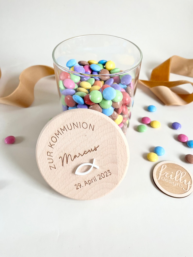 May include: A wooden jar with a lid that says "Zur Kommunion Marcus 29. April 2023" and a small white fish symbol. The jar is filled with colorful candy.