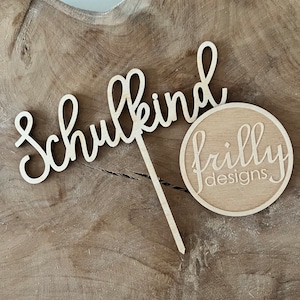 May include: A wooden cake topper with the word "Schulkind" in cursive script and a circular logo that reads "frilly designs". The topper is made of light-colored wood and is set against a wood grain background.