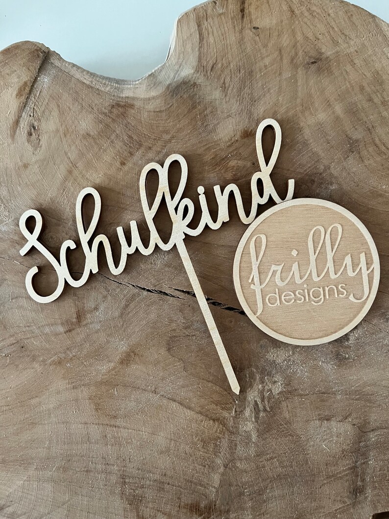 May include: A wooden cake topper with the word "Schulkind" in cursive script and a circular logo that reads "frilly designs". The topper is made of light-colored wood and is set against a wood grain background.