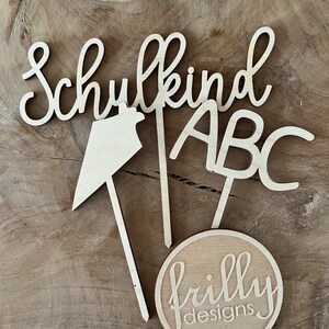 May include: Wooden cutouts spelling "Schulkind" and "ABC", along with a directional arrow and a circular logo that reads "frilly designs". The items are made of light-colored wood and are arranged on a wooden surface.