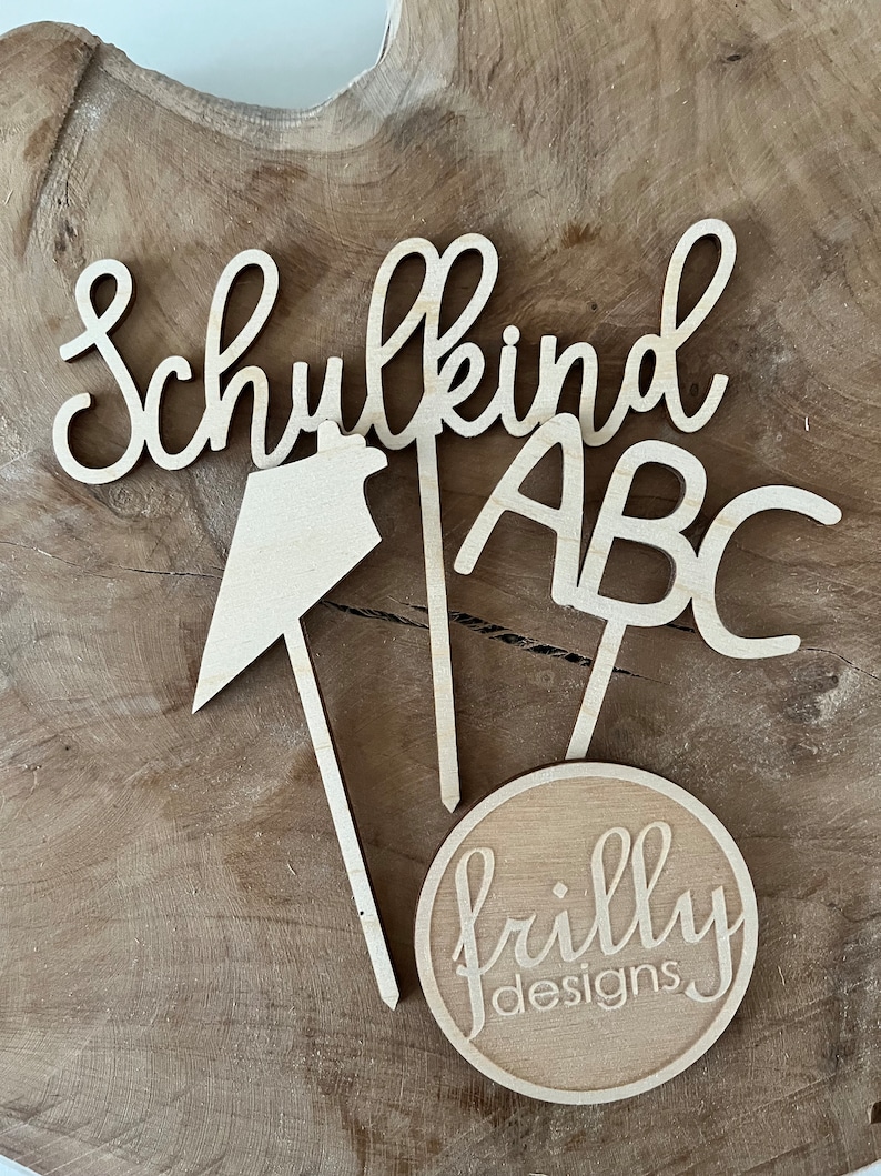 May include: Wooden cutouts spelling "Schulkind" and "ABC", along with a directional arrow and a circular logo that reads "frilly designs". The items are made of light-colored wood and are arranged on a wooden surface.