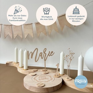 May include: A wooden birthday ring with a circular base, a name topper reading Marie, a number one topper, white candles, and a small glass vase. Above, a wooden pennant banner hangs with three circular icons featuring German text.