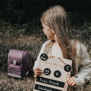 May include: A wooden sign with black chalkboard inserts for a child's first day of school. The sign says "My first day of school" and includes the date, August 1st, the child's height, 125 cm, the grade, 1st, and the teacher's name, Frau Haack. The child's desired profession is listed as "Erzieherin".