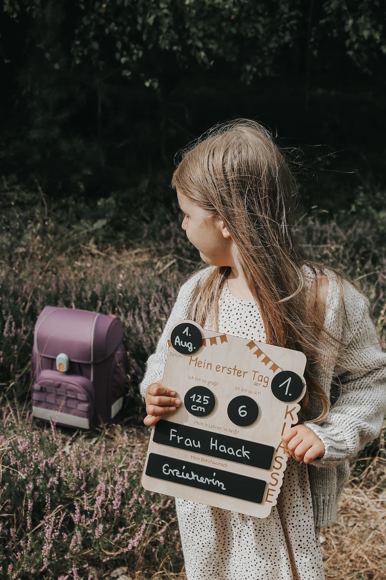 May include: A wooden sign with black chalkboard inserts for a child's first day of school. The sign says "My first day of school" and includes the date, August 1st, the child's height, 125 cm, the grade, 1st, and the teacher's name, Frau Haack. The child's desired profession is listed as "Erzieherin".