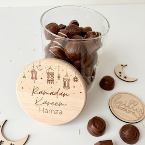 May include: A wooden jar with a lid that says "Ramadan Kareem Hamza" and has a design of crescent moons and lanterns. The jar is filled with chocolate candies.
