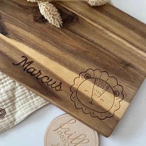 May include: Personalized wooden cutting board with the name "Marcus" engraved on it. The board features a cute lion design and a small round wooden tag with the text "frilly designs" engraved on it.