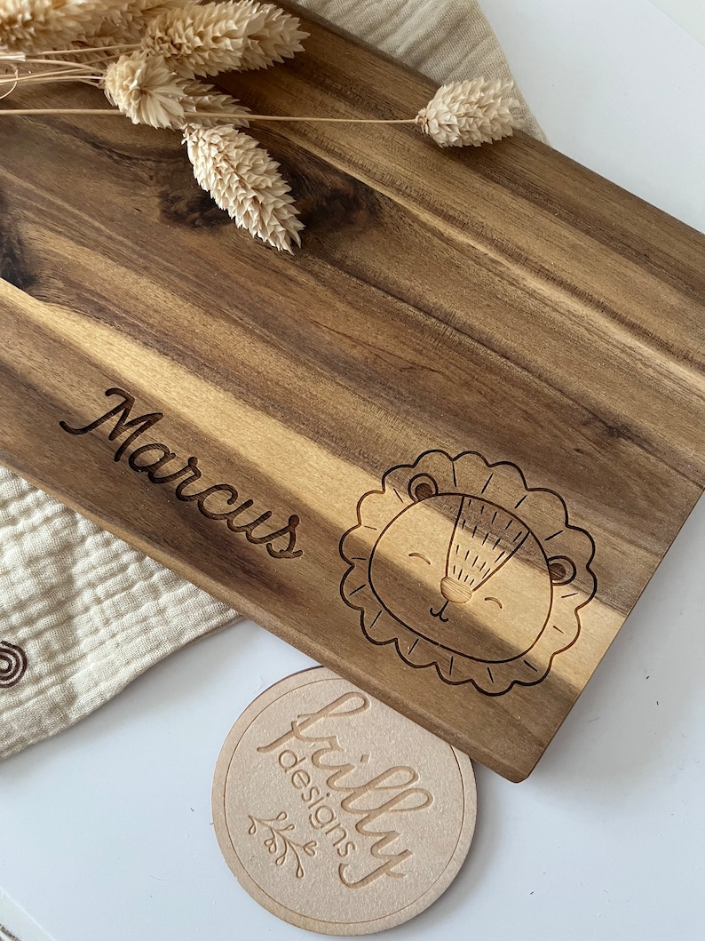 May include: Personalized wooden cutting board with the name "Marcus" engraved on it. The board features a cute lion design and a small round wooden tag with the text "frilly designs" engraved on it.