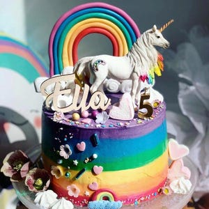 Könnte beinhalten: Ein Regenbogenkuchen, verziert mit Streuseln, einer Einhornfigur und einem Holzschild mit der Aufschrift "Ella".