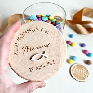 May include: A wooden lid for a jar with the text "Zur Kommunion Marcus 29. April 2023" and a small fish symbol.