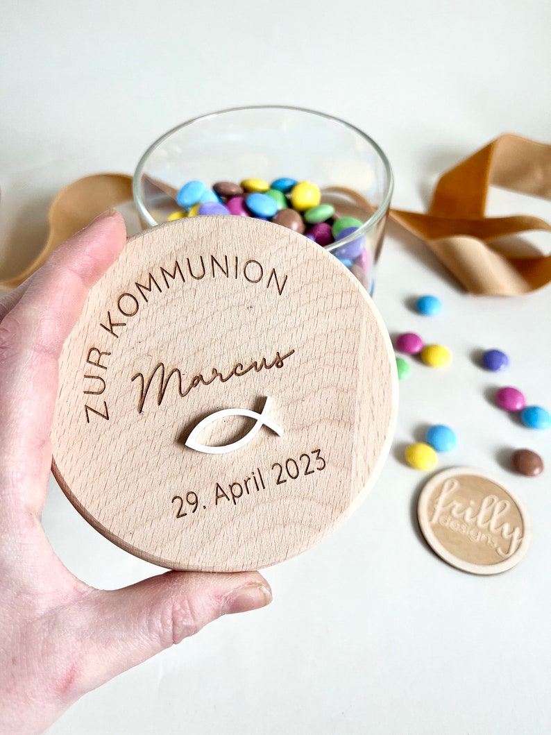 May include: A wooden lid for a jar with the text "Zur Kommunion Marcus 29. April 2023" and a small fish symbol.