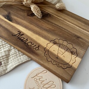 May include: A wooden cutting board with the name "Marcus" engraved on it. A cute lion face is also engraved on the board. A small round wooden tag with the text "frilly designs" is in the foreground.