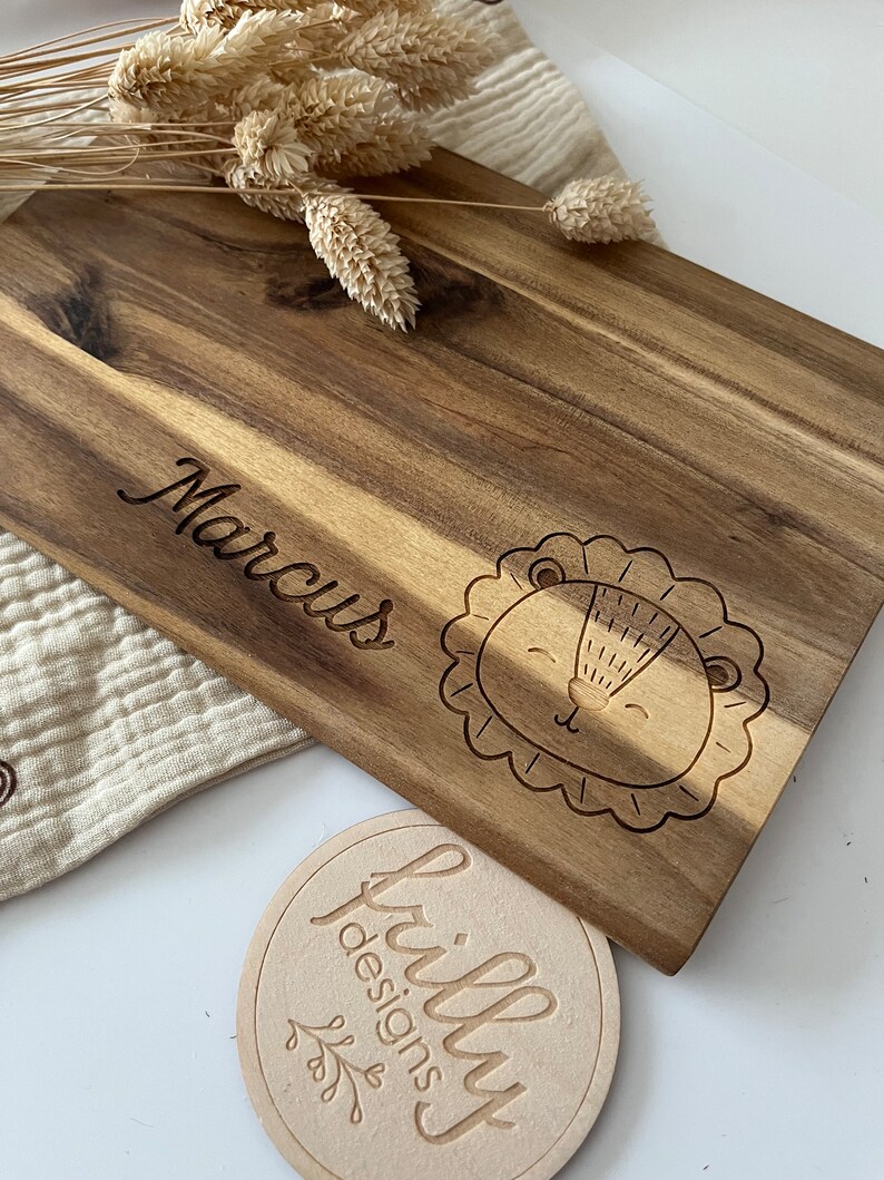 May include: A wooden cutting board with the name "Marcus" engraved on it. A cute lion face is also engraved on the board. A small round wooden tag with the text "frilly designs" is in the foreground.