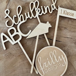 May include: Wooden laser-cut decorations with the words "Schulkind", "ABC", and "1. Klasse". A round wooden piece with the text "frilly designs" is also included. The decorations are light brown and are arranged on a wooden surface.