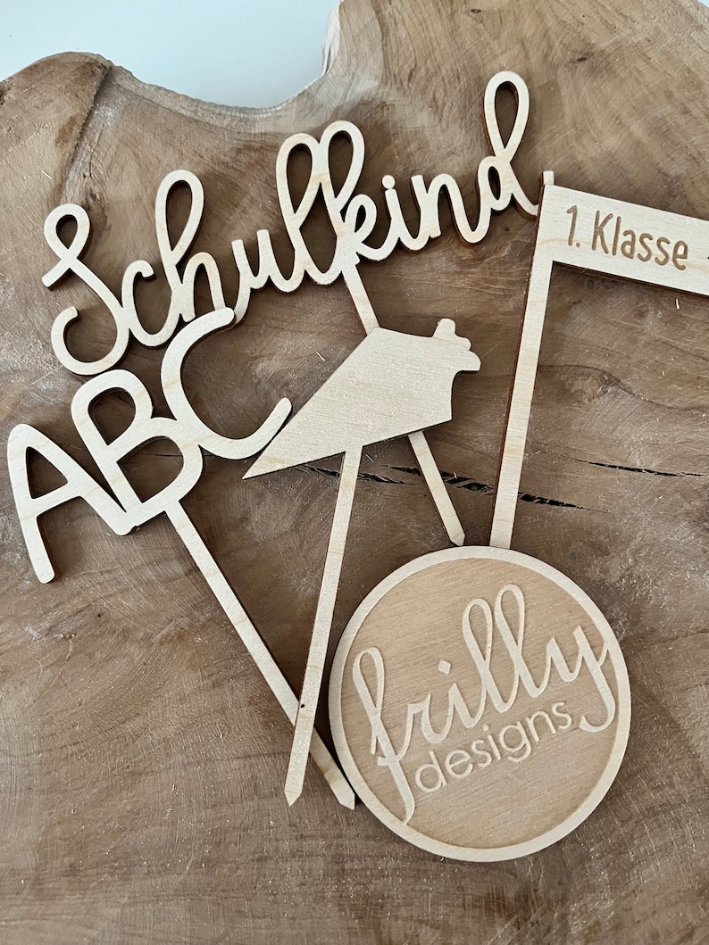 May include: Wooden laser-cut decorations with the words "Schulkind", "ABC", and "1. Klasse". A round wooden piece with the text "frilly designs" is also included. The decorations are light brown and are arranged on a wooden surface.