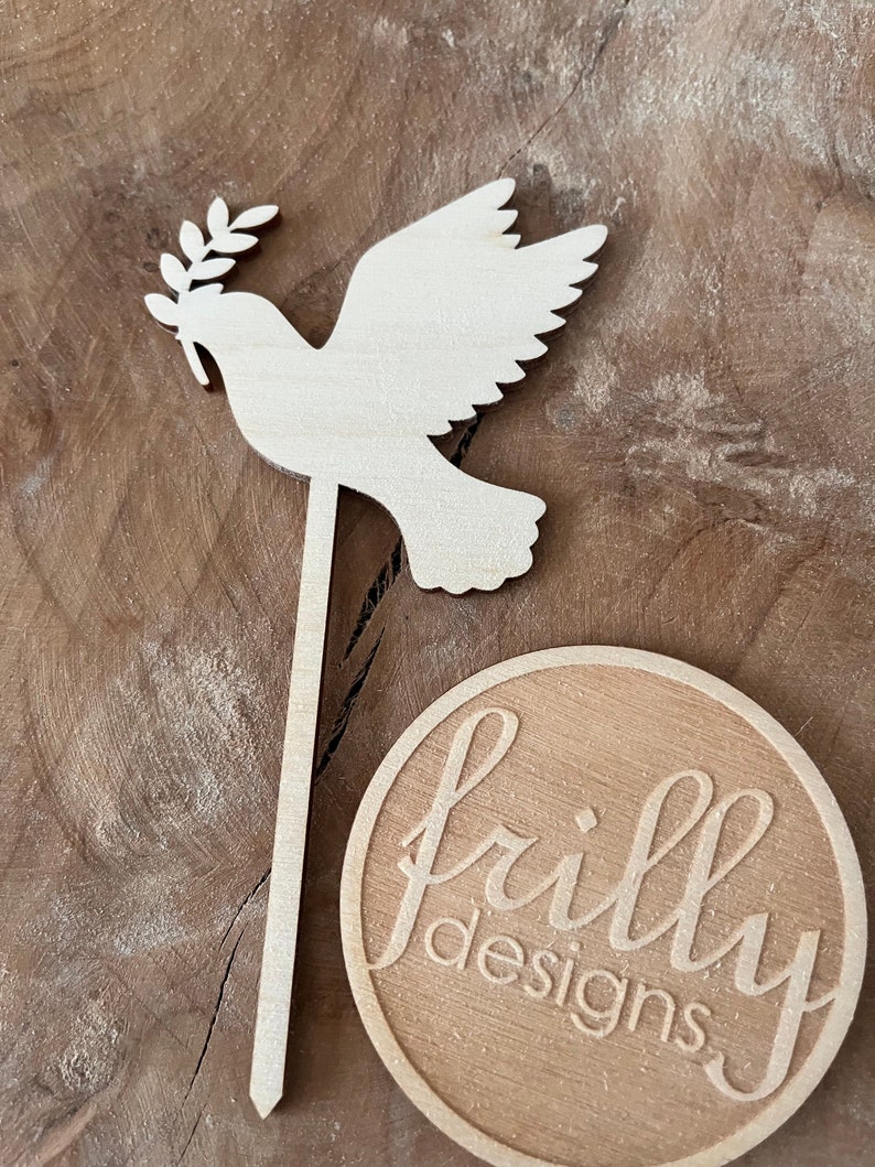 May include: Wooden dove with an olive branch in its beak, on a stick, with a round wooden sign that says "frilly designs".