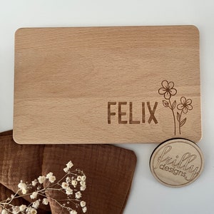 Peut inclure: Une planche &agrave; d&eacute;couper rectangulaire en bois avec des coins arrondis. Le nom "FELIX" est grav&eacute; sur la planche, ainsi qu'un motif floral. Un petit dessous de verre rond en bois avec le logo "frilly designs" est &eacute;galement pr&eacute;sent.