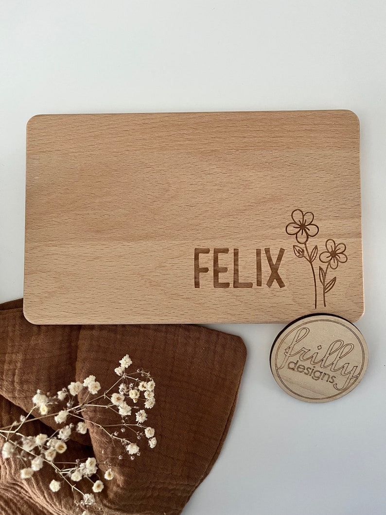 Peut inclure: Une planche &agrave; d&eacute;couper rectangulaire en bois avec des coins arrondis. Le nom "FELIX" est grav&eacute; sur la planche, ainsi qu'un motif floral. Un petit dessous de verre rond en bois avec le logo "frilly designs" est &eacute;galement pr&eacute;sent.