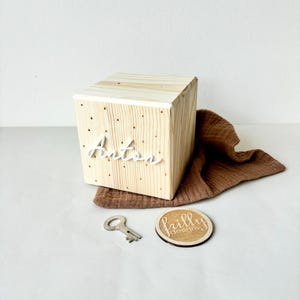 May include: A wooden box with the name Anton written on the front in white lettering. The box has a keyhole and is sitting on a brown fabric. A small wooden circle with the text "frilly designs" is next to the box.