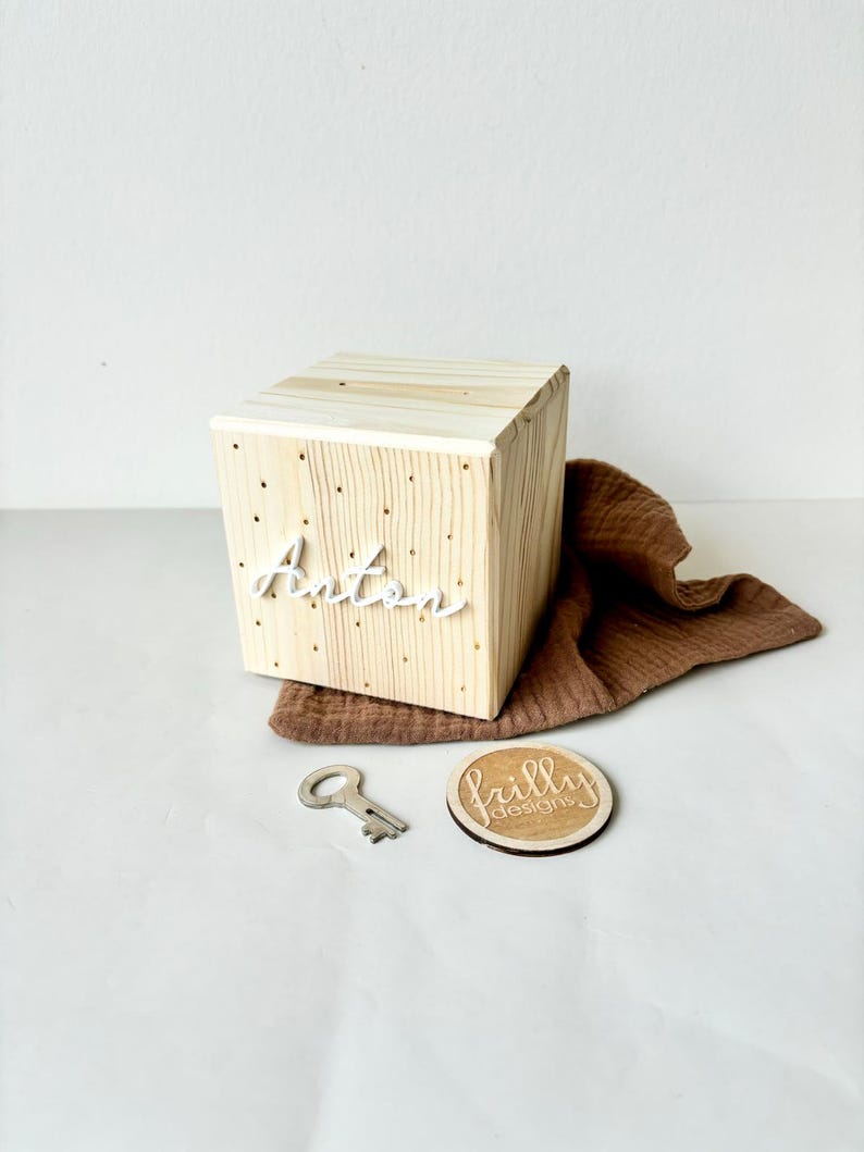 May include: A wooden box with the name Anton written on the front in white lettering. The box has a keyhole and is sitting on a brown fabric. A small wooden circle with the text "frilly designs" is next to the box.
