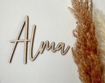 Puede incluir: Un letrero de madera con el nombre "Alma" escrito en cursiva. El letrero es de madera marrón claro y está sobre un fondo blanco.