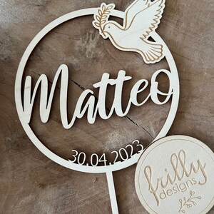 May include: Wooden cake topper with a circle design featuring the name Matteo and the date 30.04.2023. A dove with an olive branch is in the circle. The topper is on a wooden stick. A round wooden tag with the text "frilly designs" is in the lower right corner.