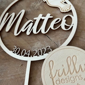 May include: Wooden cake topper with a circle design featuring the name Matteo, the date 30.04.2023, and a dove with an olive branch. The topper is attached to a wooden stick. A separate wooden circle with the text "frilly designs" is also shown.
