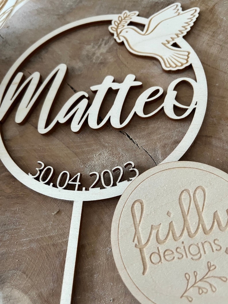 May include: Wooden cake topper with a circle design featuring the name Matteo, the date 30.04.2023, and a dove with an olive branch. The topper is attached to a wooden stick. A separate wooden circle with the text "frilly designs" is also shown.