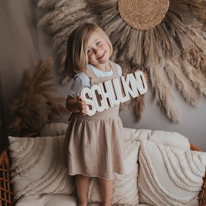 May include: A young girl in a beige dress stands on a couch holding a wooden sign that says "SCHULKIND". The sign is white with black lettering. The girl is smiling and looking at the camera. The couch is brown wicker with white cushions.