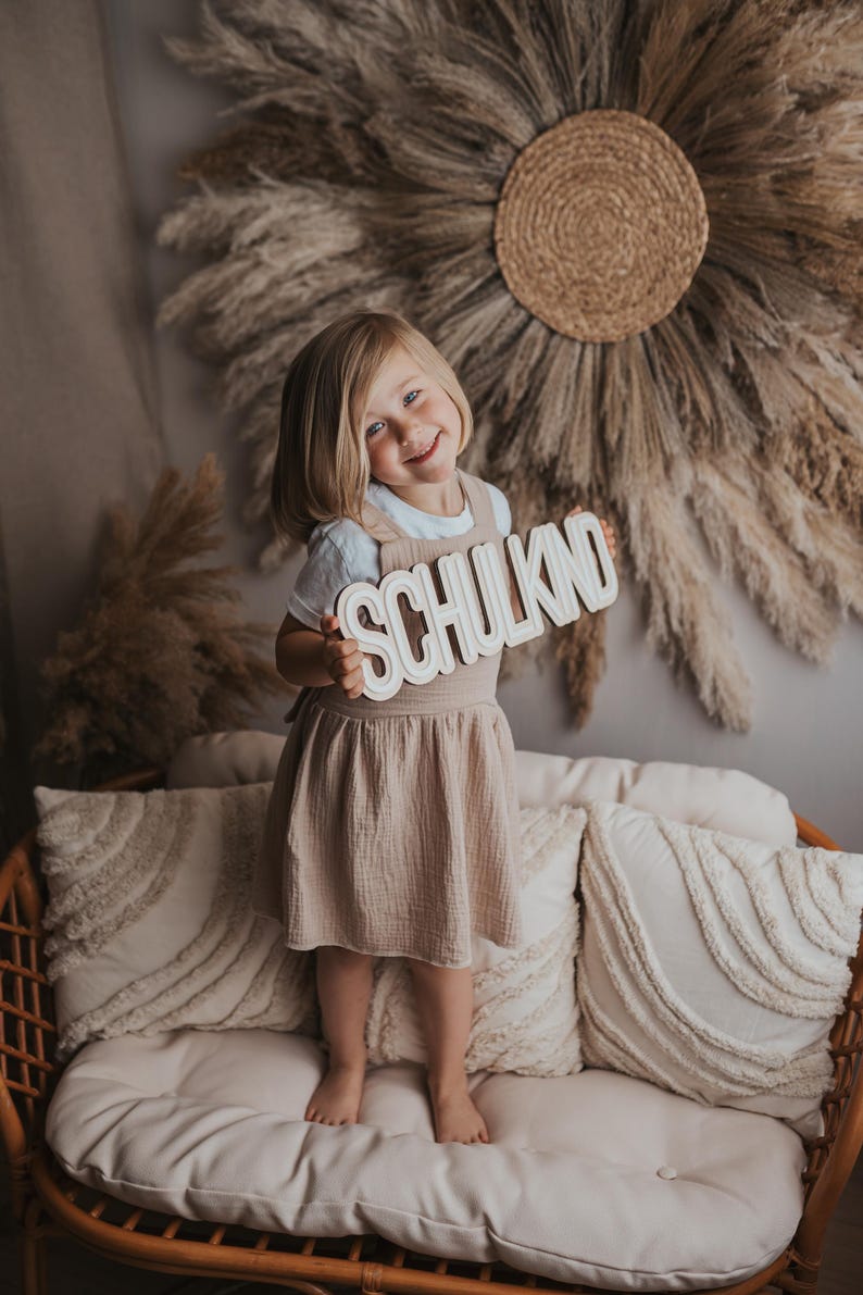 May include: A young girl in a beige dress stands on a couch holding a wooden sign that says "SCHULKIND". The sign is white with black lettering. The girl is smiling and looking at the camera. The couch is brown wicker with white cushions.