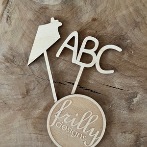 May include: Wooden cake toppers with the letters "ABC", a house-shaped design, and a circular design with the text "frilly designs" in a cursive font. The toppers are made of light-colored wood.