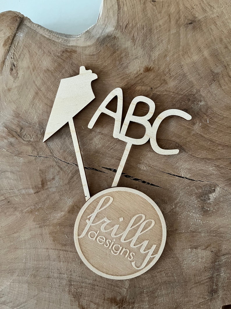 May include: Wooden cake toppers with the letters "ABC", a house-shaped design, and a circular design with the text "frilly designs" in a cursive font. The toppers are made of light-colored wood.