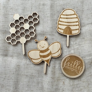 Könnte beinhalten: Vier hölzerne, lasergeschnittene Kuchentopper zum Thema Bienen. Die Topper beinhalten eine Wabe, eine Biene, einen Bienenstock und einen runden Topper mit dem Text "frilly designs".