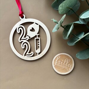 Op de afbeelding: Een houten kerstversiering met het jaartal 2022, een spuit en een hartvormig ontwerp. De ronde ornament is opgehangen met een rood lint. Een kleine houten cirkel met de tekst "frilly designs" is ook zichtbaar.