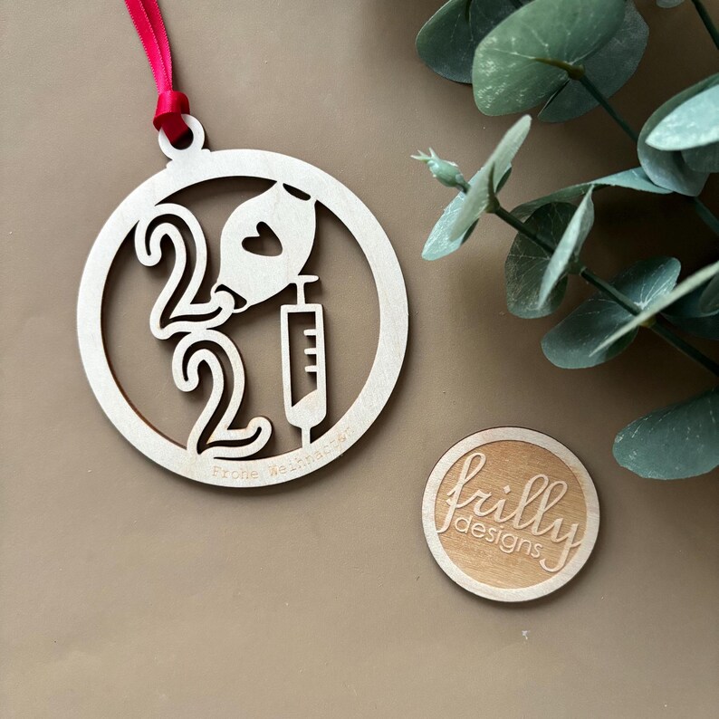 Op de afbeelding: Een houten kerstversiering met het jaartal 2022, een spuit en een hartvormig ontwerp. De ronde ornament is opgehangen met een rood lint. Een kleine houten cirkel met de tekst "frilly designs" is ook zichtbaar.