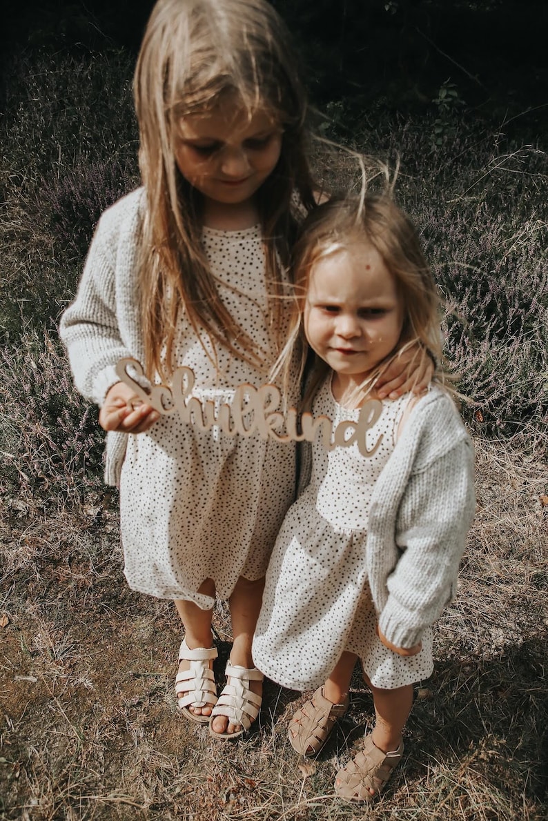 May include: Two young girls wearing white dresses with black polka dots and beige cardigans are holding a wooden sign that says "Schulkind". They are standing in a field of green grass and purple flowers.