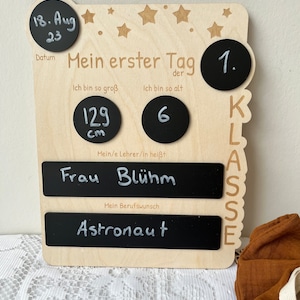 May include: Wooden first day of school sign with chalkboard elements. The sign includes a date, height, age, teacher's name, and a space for a future career. The sign is decorated with stars and the words "Mein erster Tag" (My first day).