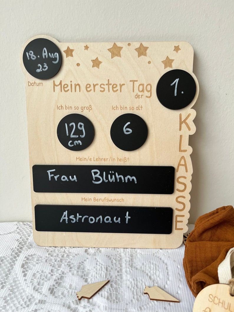 May include: Wooden first day of school sign with chalkboard elements. The sign includes a date, height, age, teacher's name, and a space for a future career. The sign is decorated with stars and the words "Mein erster Tag" (My first day).