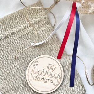 Op de afbeelding: Een houten rond bord met de tekst "frilly designs" ligt op een jute zak. Rode, witte en blauwe linten zijn bevestigd. Gedroogde tarwestengels en touw zijn ook zichtbaar.