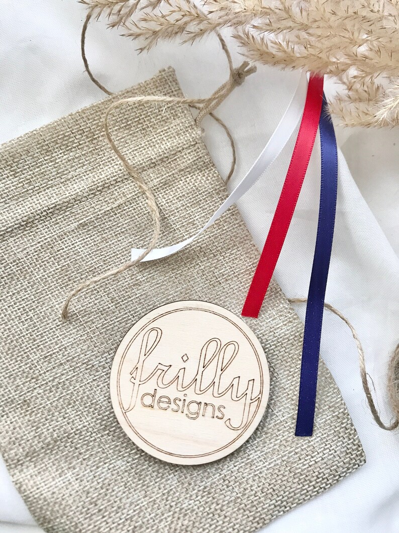 Op de afbeelding: Een houten rond bord met de tekst "frilly designs" ligt op een jute zak. Rode, witte en blauwe linten zijn bevestigd. Gedroogde tarwestengels en touw zijn ook zichtbaar.