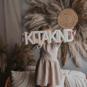 May include: A young child holding a wooden sign that says "KITAKIND" in white letters. The child is wearing a light brown dress and is standing in front of a wall decorated with dried pampas grass.