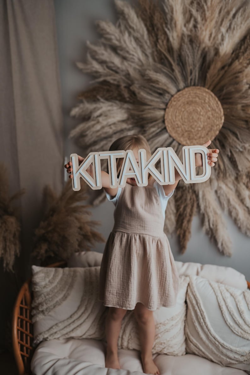 May include: A young child holding a wooden sign that says "KITAKIND" in white letters. The child is wearing a light brown dress and is standing in front of a wall decorated with dried pampas grass.