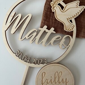 May include: Wooden circle with the name Matteo and the date 30.04.2023 engraved. A dove with an olive branch is on the right side of the circle. A smaller circle with the text "frilly designs" is below the larger circle.