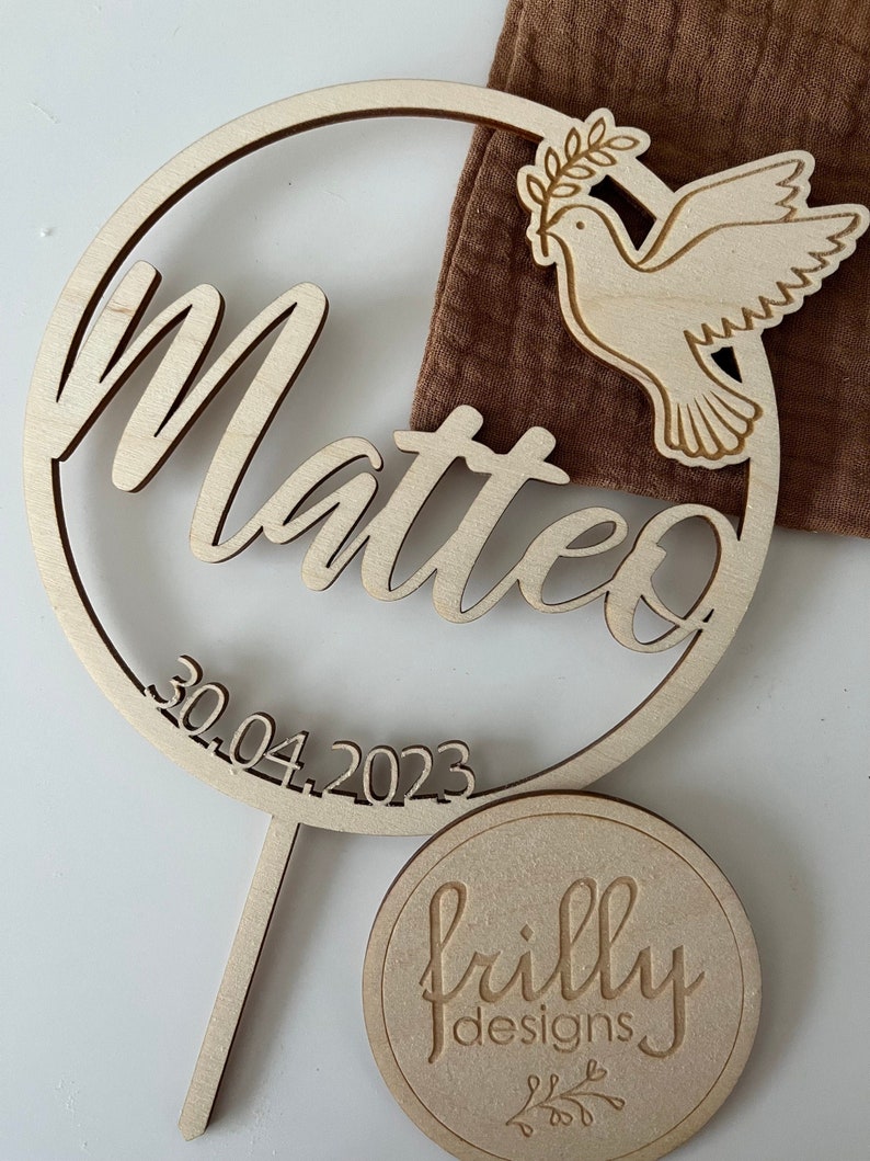 May include: Wooden circle with the name Matteo and the date 30.04.2023 engraved. A dove with an olive branch is on the right side of the circle. A smaller circle with the text "frilly designs" is below the larger circle.