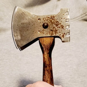 May include: A vintage hatchet with a wooden handle and a metal head. The head features a blade and a toothed edge. The handle is a dark brown color. The metal head has some rust.