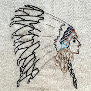 May include: Embroidered Native American headdress design in black, tan, blue, and brown thread. The profile of a person wearing a feathered headdress is stitched on a tan fabric background with a subtle herringbone pattern.