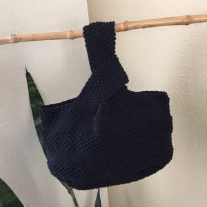 May include: A navy blue knitted tote bag with a textured pattern. The bag has a single handle and is suspended from a bamboo rod. The bag's design features a rounded bottom and a structured shape, showcasing a handmade aesthetic.