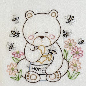 May include: Embroidered white bear with a honey pot, surrounded by bees and flowers. The bear is holding a pot of honey with the word "Honey" on it. The design includes pink, green, brown, and yellow colors.