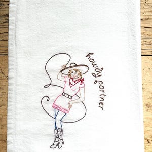 May include: White kitchen towel with an embroidered design of a cowgirl holding a lasso. The cowgirl is wearing a pink top, skirt, and boots, with a red bandana and a cowboy hat. The words "howdy partner" are written in brown.